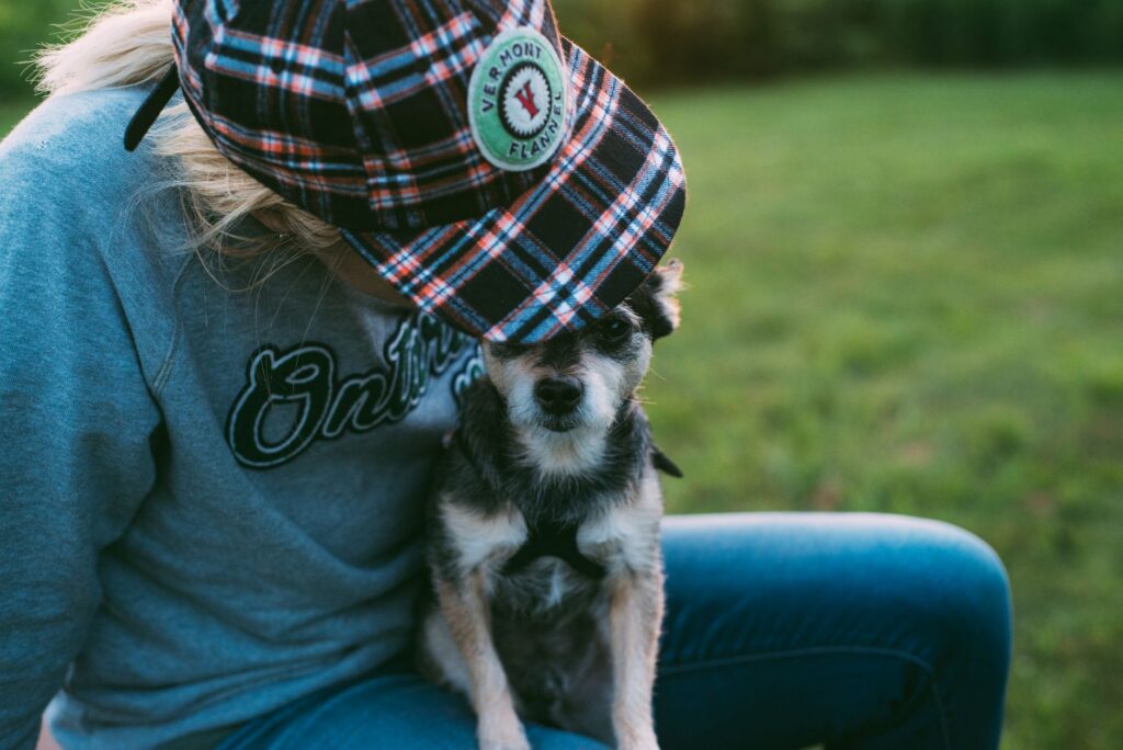 A person in a plaid cap sits with a small dog on the grass, capturing a moment of affection.