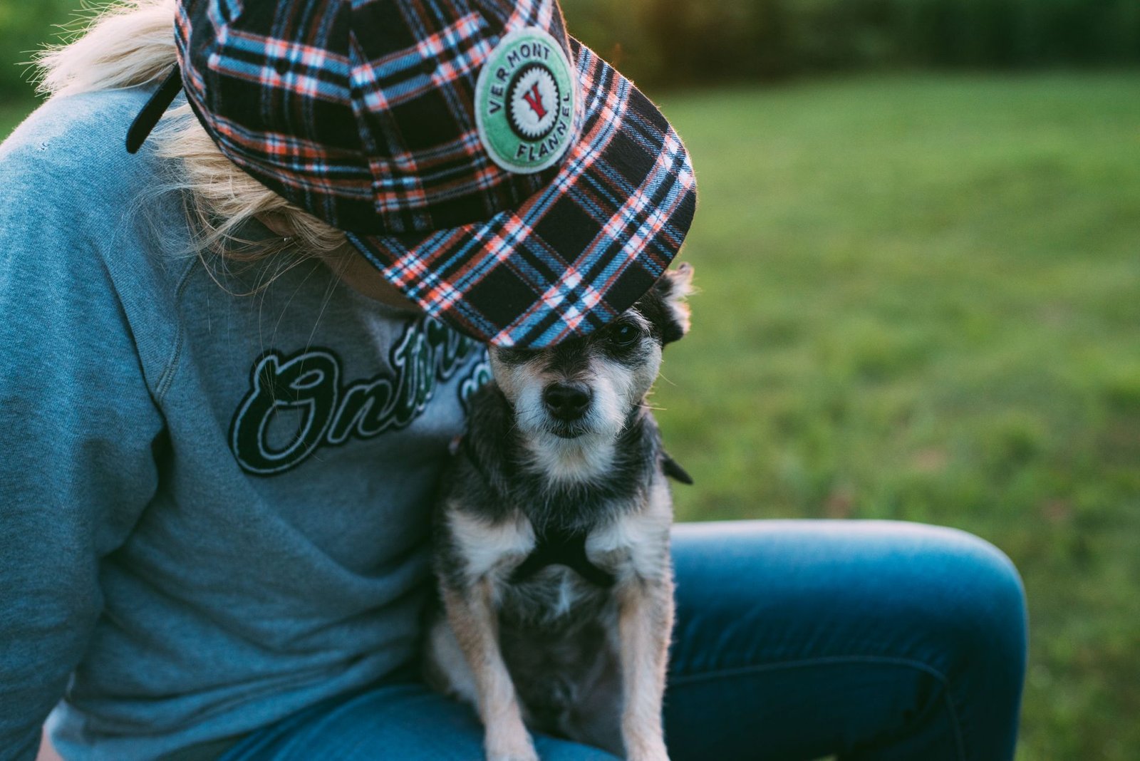 A person in a plaid cap sits with a small dog on the grass, capturing a moment of affection.